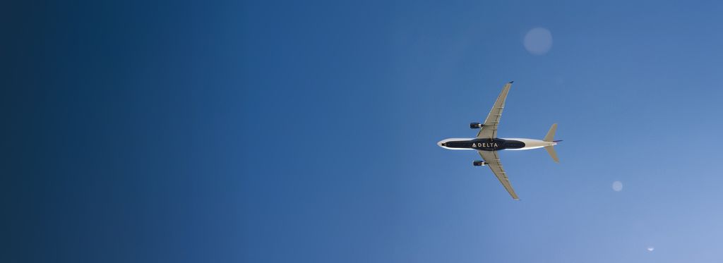 A Delta airplane flying across a clear blue sky, shown from an underside view. The word “DELTA” appears in large, white lettering on the bottom of the aircraft against the dark blue background.