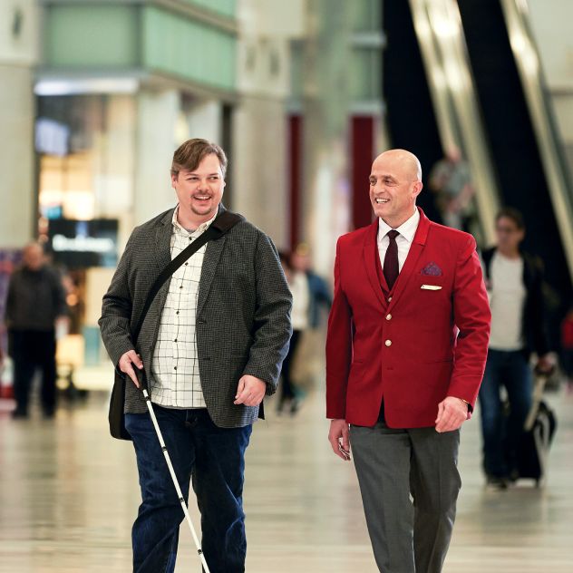A Delta employee in uniform walks alongside a traveler using a white cane through the airport concourse, with other travelers visible in the background.
