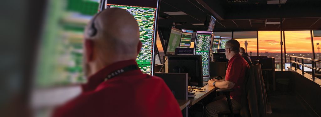 A Delta employee sits at a workstation in an operations control center, facing multiple computer monitors filled with data and maps. Large windows in the background reveal a sunset sky and airport structures in the distance.