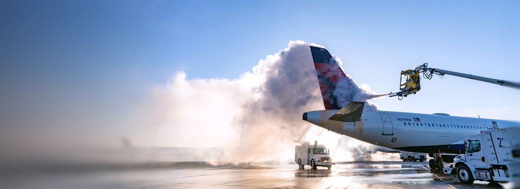 A Delta airplane on the tarmac is being deiced, with fluid spraying over the tail from an elevated boom truck and creating a cloud of mist against a clear blue sky.