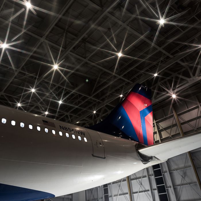 A close-up of a Delta airplane’s red and blue tail section inside a hangar. Overhead lights and structural beams of the hangar ceiling are clearly visible above the aircraft.