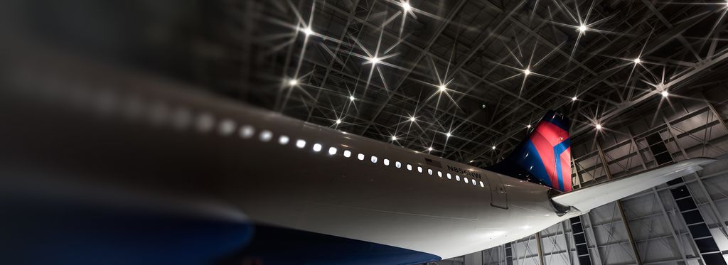 A close-up of a Delta airplane’s red and blue tail section inside a hangar. Overhead lights and structural beams of the hangar ceiling are clearly visible above the aircraft.