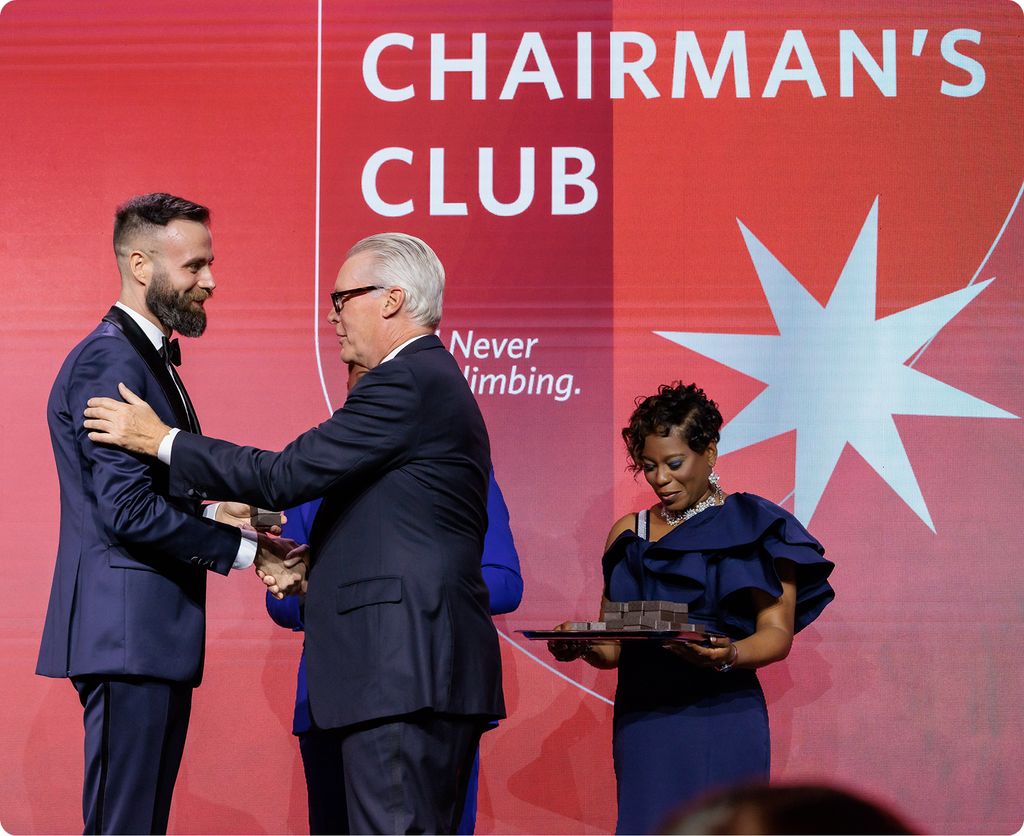 Delta CEO, Ed Bastian, shakes the hand of an employee on a stage with a red backdrop reading “Chairman’s Club”. A third person stands nearby holding a tray of awards.