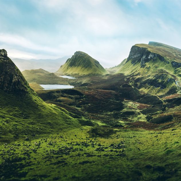 A wide view of a lush green valley with rolling hills, winding paths, and steep mountains rising in the background. Patches of sunlight highlight the landscape under a pale sky, creating a panoramic, natural scene.