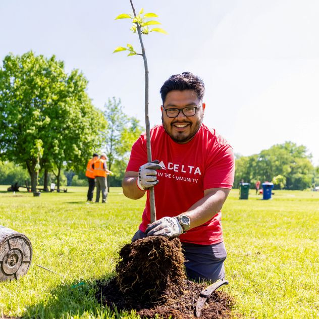 A Delta employee volunteer wearing a red “Delta in the Community” T‑shirt and work gloves kneels on grass while holding a plant, preparing to place it in the ground. Other volunteers and trees are visible in the background.