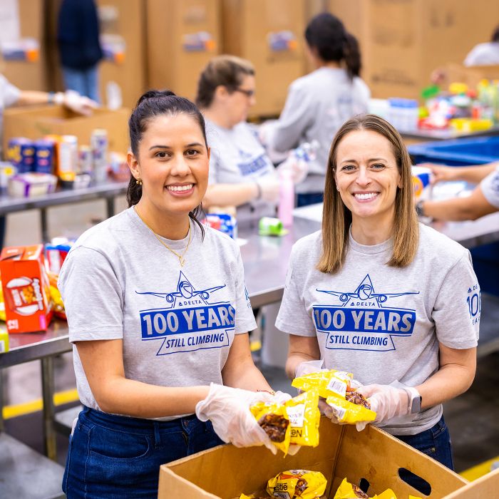 Two Delta employees stand side by side wearing matching gray T‑shirts that read “100 YEARS – STILL CLIMBING.” They hold packaged food items, with tables, boxes, and other volunteers working in the background