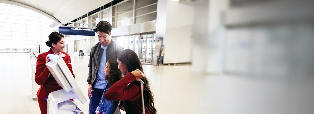 A Delta employee in a red uniform assists a family of three at a self‑service kiosk in a bright, modern airport concourse.