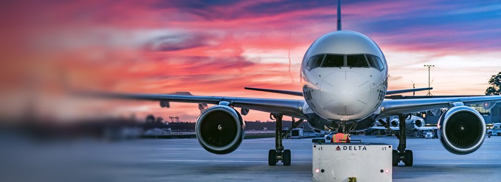 A close up, front-facing  view of a Delta airplane on the tarmac at sunset, with an employee in an orange high‑visibility vest standing directly in front of the nose and facing the aircraft. Pink and orange clouds fill the sky, with airport equipment visible beneath the wings.