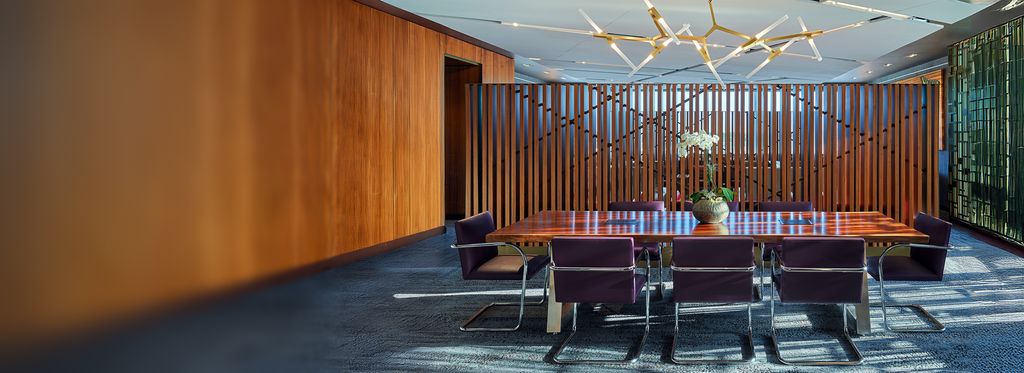 A modern conference room with a long wooden table surrounded by purple upholstered chairs. A potted plant sits at the center of the table, with vertical wood slats and glass walls in the background.