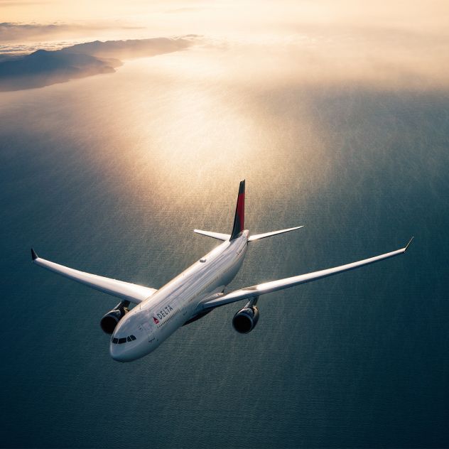 A Delta airplane flies above open water, viewed from above, with sunlight reflecting off the wings and the ocean below glowing in warm light.