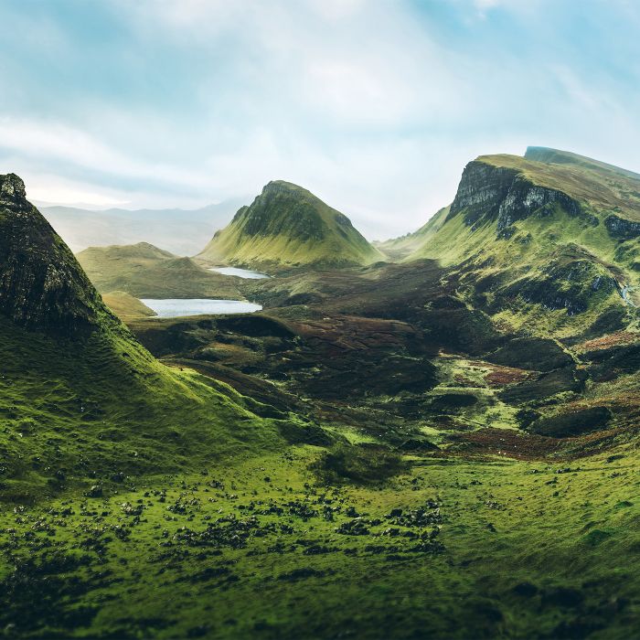 A wide view of a lush green valley with rolling hills, winding paths, and steep mountains rising in the background. Patches of sunlight highlight the landscape under a pale sky, creating a panoramic, natural scene.