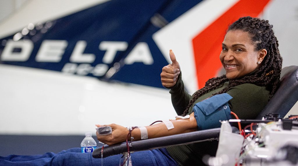 A blood donor reclines in a donation chair with a blood pressure cuff and tubing attached to their arm, giving a thumbs‑up. A blurred Delta airplane tail is visible in the background