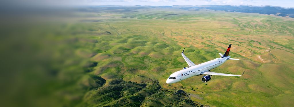 A Delta airplane flies above a green landscape of fields and rolling terrain, viewed from an elevated angle, with the aircraft’s white body and red‑and‑blue tail clearly visible