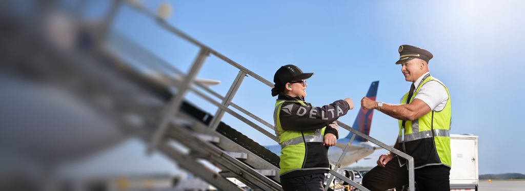 A Delta pilot and mechanic wearing high‑visibility vests exchange a fist bump outdoors at the aircraft boarding stairs beside an airplane under a clear blue sky.