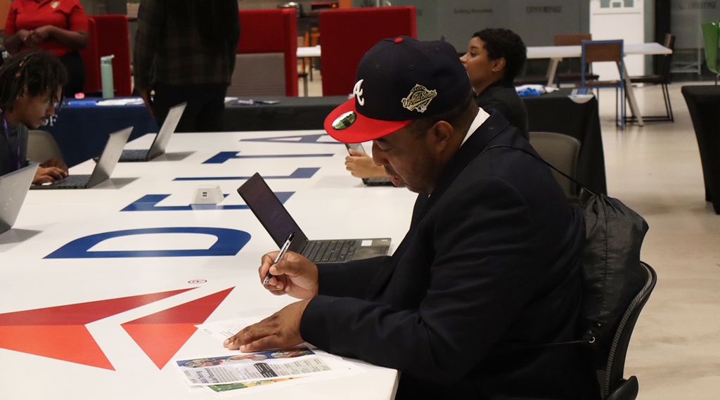 A person sits at a table signing a large white surface with the Delta logo printed in blue and red. They wear a dark jacket and a black and red baseball cap, with a laptop open near them. Other tables, chairs, and people are visible in the background.